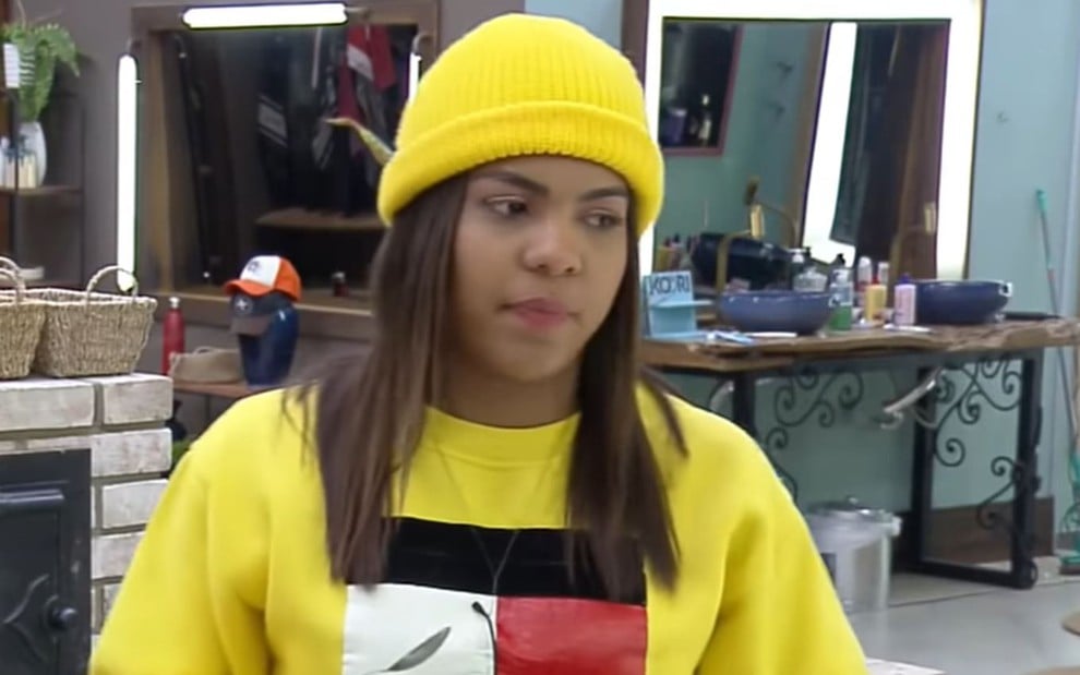 Tays está na cozinha da sede, ela usa um moletom amarelo com detalhes preto, branco e vermelho e uma touca amarela
