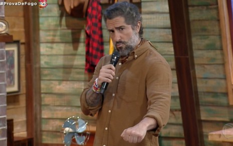 Marcos Mion fazendo bico, com uma camisa marrom e segurando um microfone