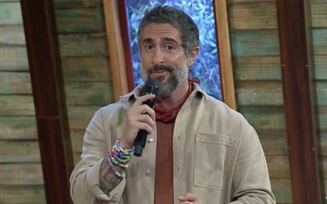 Marcos Mion segurando o microfone, vestindo uma jaqueta clara