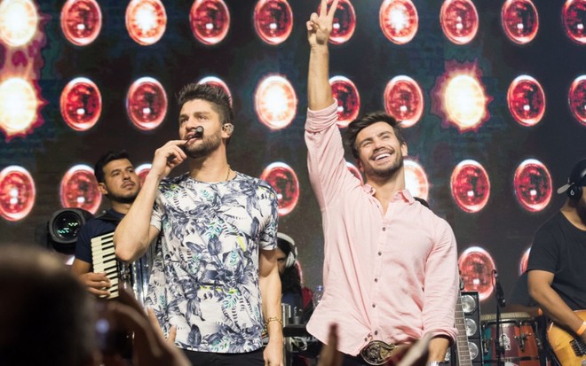 Imagem de Munhoz e Mariano em fundo preto e vermelho, durante uma apresentação musical.