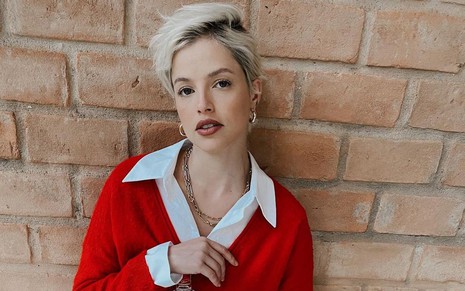 A atriz Agatha Moreira, com cabelo curto e loiro, posa para campanha publicitária com camisa branca e casaco vermelho
