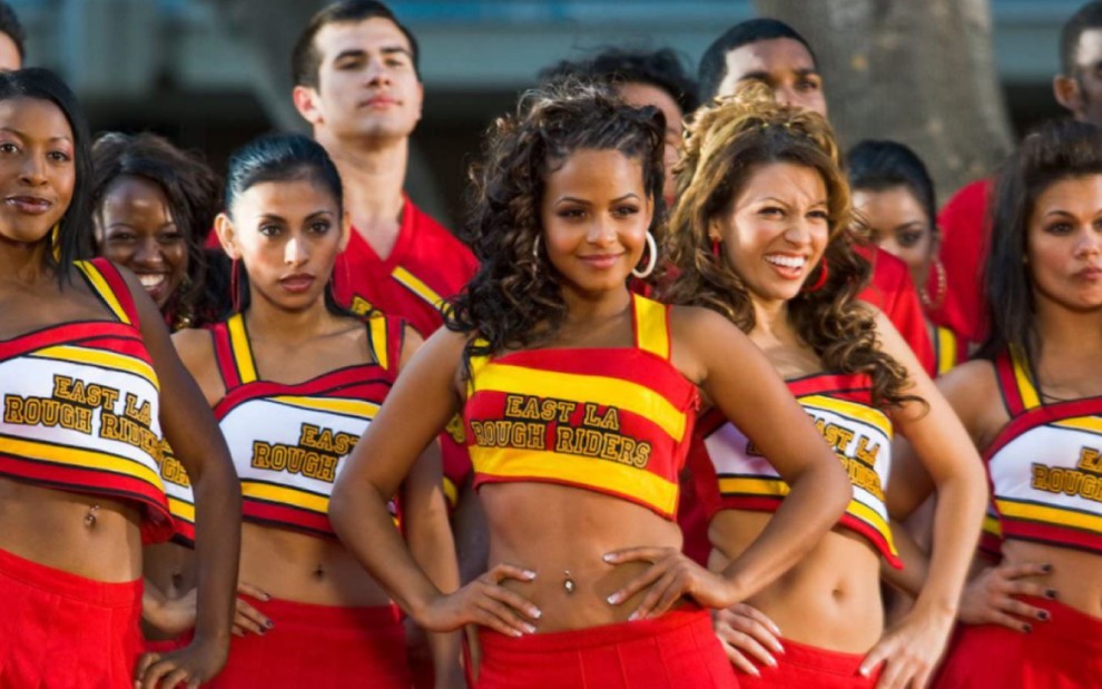 Um grupo de meninas à frente, usando roupas de líderes de torcida, vermelho, amarelo e branco