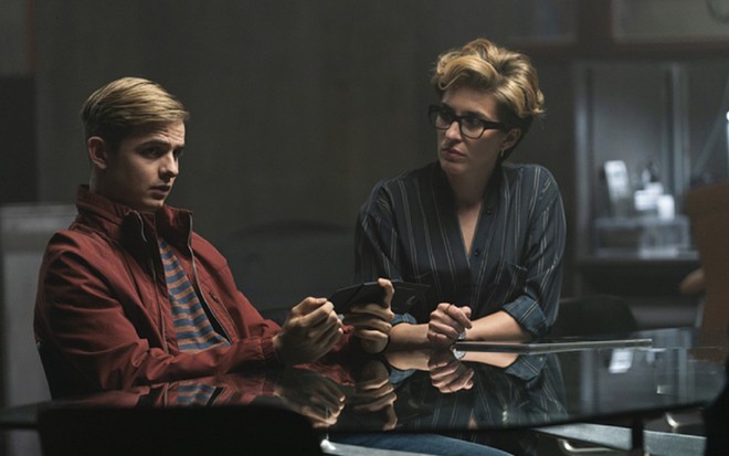 Cena do seriado Alex Rider mostra os atores Otto Farrant e  Vicky McClure sentados lado a lado à frente de uma mesa de vidro. Ele é jovem, tem o cabelo penteado para o lado e usa jaqueta vermelha. Ela tem 37 anos, usa óculos e uma camisa social escura