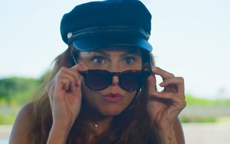 A atriz Deborah Secco usa um óculos escuro e uma boina preta em cena como Alexia/Josimara de Salve-se Quem Puder