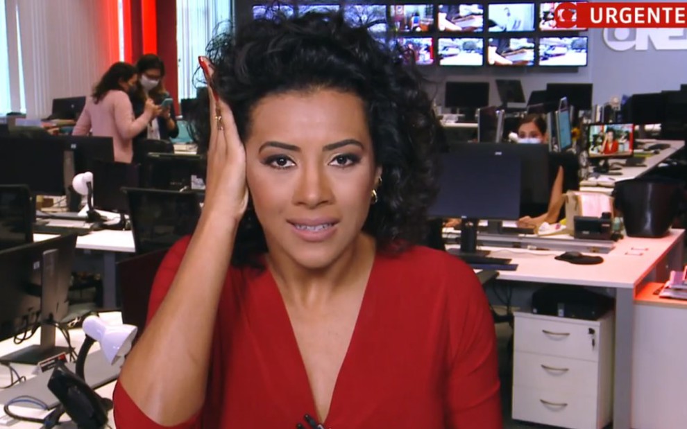A jornalista Aline Midlej com uma das mãos na cabeça e vestindo uma blusa decotada vermelha