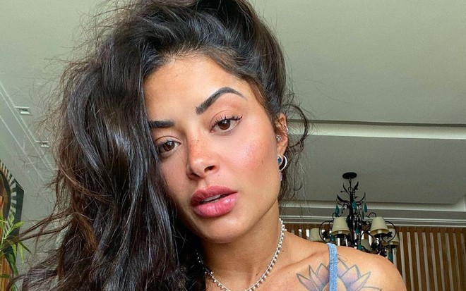 Aline Riscado olha para câmera, está com o cabelo (castanho) jogado para o lado e blusa de alça azul