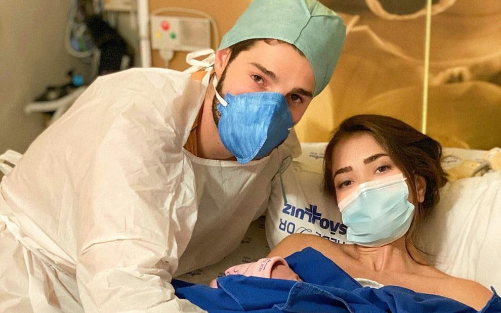 Alok está com roupa de hospital e máscara ao lado de Romana, que está deitada na cama de hospital e também está de máscara; ela segura o bebê