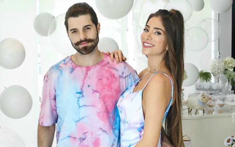 Alok está ao lado de Romana; ele usa camiseta colorida (azul e rosa); ela está com o cabelo solto e usa vestido de alça nas cores azul e rosa