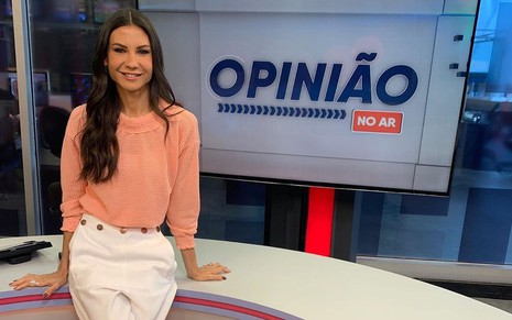 Amanda Klein de calça branca e blusa rosé sentada na bancada do Opinião no Ar, da RedeTV!
