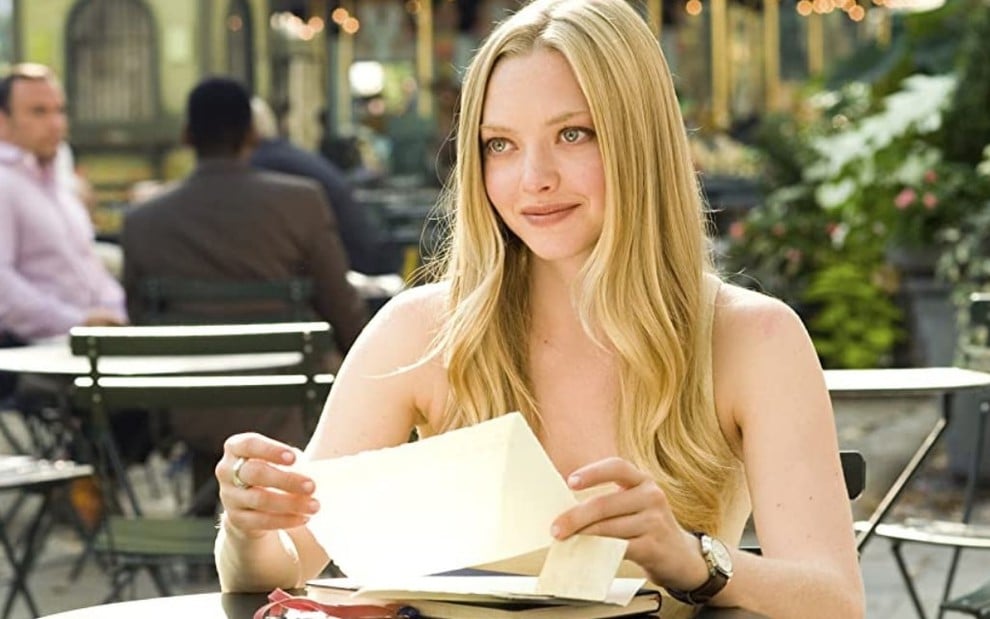 Amanda Seyfried sentada em uma mesa num restaurante ao ar livre; ela segura nas mãos uma carta em cena do filme Cartas para Julieta (2010)