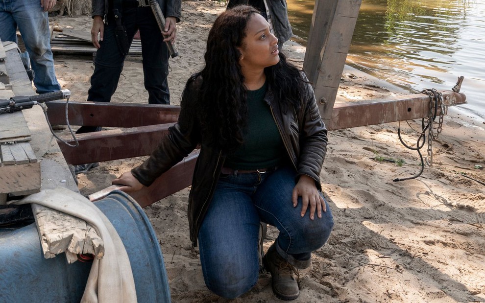Agachada ao lado de um barco, a atriz Aliyah Royale olha para o lado em cena de The Walking Dead: World Beyond