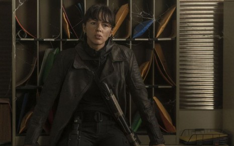 Annet Mahendru, com roupas escuras e uma grande arma, se escora em uma estante em cena de The Walking Dead: World Beyond