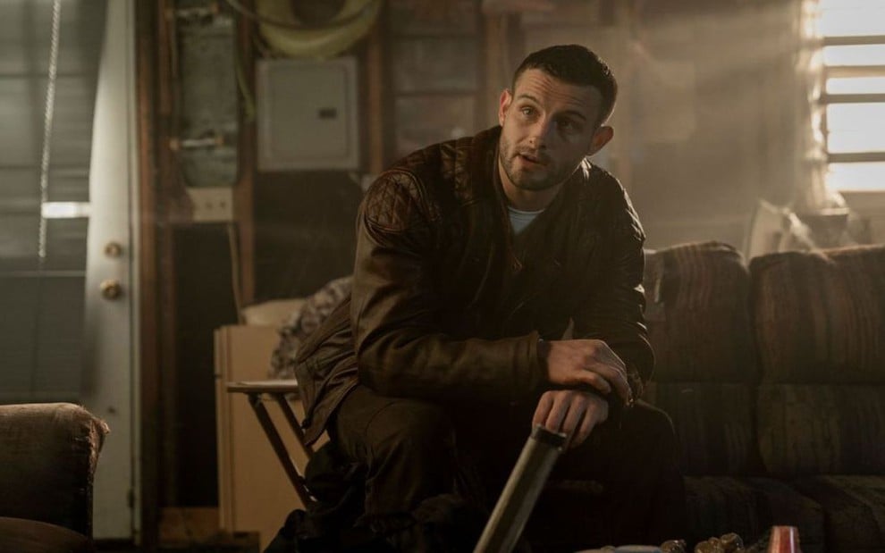 Sentado em um sofá antigo com uma jaqueta de couro, Nico Tortorella faz expressão de surpresa em cena de The Walking Dead: World Beyond