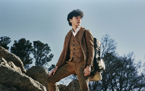 Com terno e mochila de explorador, Nicolas Cantu posa em cima de pedras com expressão determinada