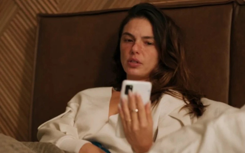 A atriz Isis Valverde deitada, faz chamada de vídeo por celular, tem rosto marcado por máscara em Amor de Mãe
