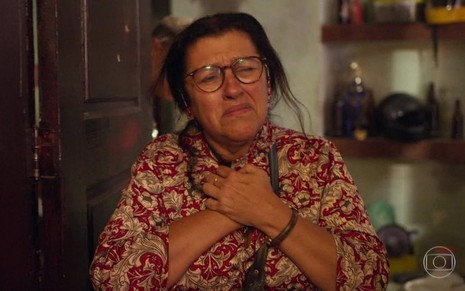 A atriz Regina Casé com camisa estampada, mãos sobrepostas no peito e expressão de tristeza em cena de Amor de Mãe