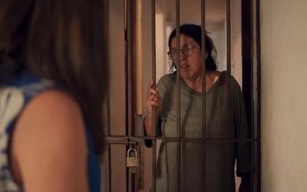 A atriz Regina Casé atrás das grades em cativeiro, conversa com Adriana Esteves, de costas, em cena de Amor de Mãe