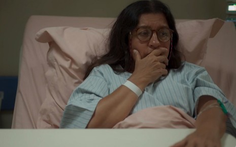 Pensativa, Regina Casé coloca a mão na boca deitada em um leito hospitalar na novela Amor de Mãe