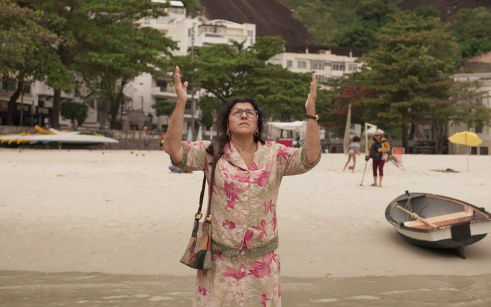 A atriz Regina Casé como Lurdes ergue os braços para o céu em oração em uma praia em cena de Amor de Mãe