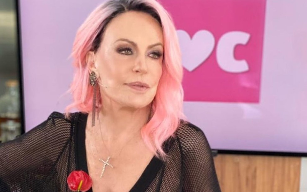 A apresentadora Ana Maria Braga com cabelo rosa, olha para a direita, com logo do Mais Você em telão ao fundo