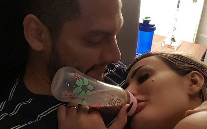 Thiago Lopes olhando Andressa Urach em seu colo; ela aparece tomando mamadeira com achocolatado