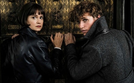 Com expressão assustada, Katherine Waterston e Eddie Redmayne aparecem em frente a uma porta em Animais Fantásticos: Os Crimes de Grindelwald (2018)