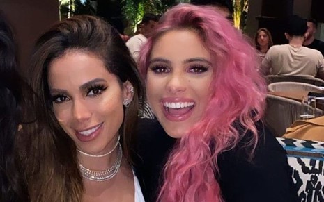 Anitta usa blusa branca com decote e está com o cabelo (castanho) solto; ao lado dela está Lele Pons, que usa casaco preto e está com o cabelo (cor de rosa) solto; as duas estão sorrindo na imagem