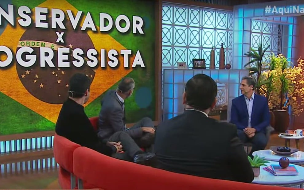 Luís Ernesto Lacombe e Nathália Batista (de frente) discutem conservadorismo no Aqui na Band de terça (23), no painel, bandeira do Brasil e as palavras 'Conservador x Progressista'