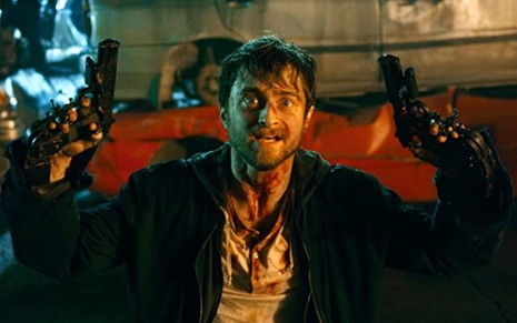 Daniel Radcliffe ajoelhado, coberto de sangue e com duas armas nas mãos em cena do filme Armas em Jogo (2019)