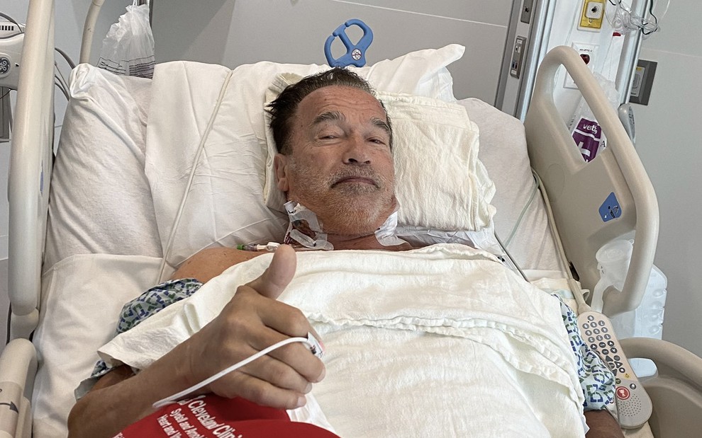 O ator Arnold Schwarzenegger em uma cama de hospital faz o sinal de "joinha" com a mão direita