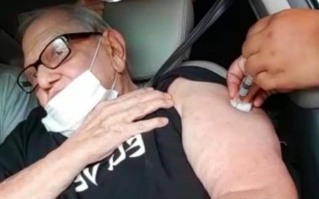 O humorista Ary Toledo levanta a manga de seu braço esquerdo para receber, de máscara, a vacina contra a covid-19 em seu carro