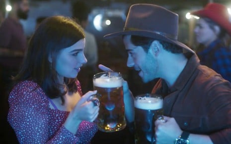 Gabriela Medvedovski e Jessé Scarpellini em cena de As Five: caracterizados como seus personagens, atriz segura caneca de cerveja e olha de maneira convidativa para ator que está com chapéu de playboy e flerta de volta