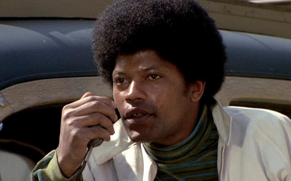 Clarence Williams 3º como Lincoln 'Linc' Hayes na série The Mod Squad com cabelo black power, camiseta verde e blusa branca segurando um rádio na mão direita