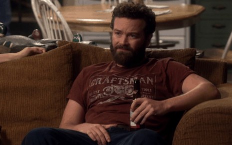 Jameson Bennett (Danny Masterson) em The Ranch sentado no sofá com camisa vermelho escuro com uma garrafa de cerveja na mão esquerda