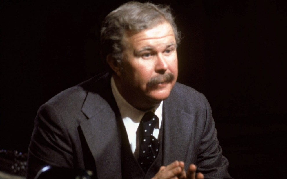 Arthur Jensen (Ned Beatty) de bigode e paletó sentado em fundo escuro olhando para o lado direito em cena do filme Rede de Intrigas (1976)