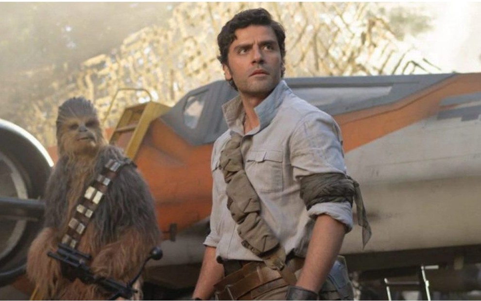 Poe Cameron (Oscar Isaac) no centro de camisa cinza com o rosto virado para a esquerda e o personagem Chewbacca no lado esquerdo com uma nave ao fundo