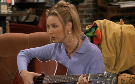 Phoebe (Lisa Kudrow) de blusa roxa sentada em um sofá e segurando um violão