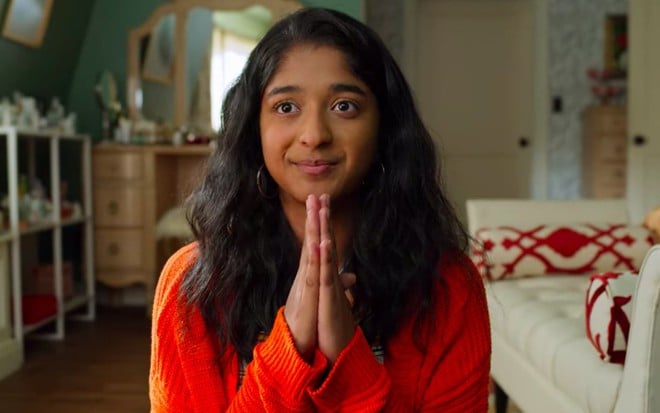 Devi (Maitreyi Ramakrishnan) de blusa laranja e as duas mãos juntas em pose de oração