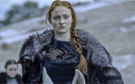 Sansa Stark (Sophie Turner) em cena de Game of Thrones com blusa azul e casaco em fundo claro com montanhas