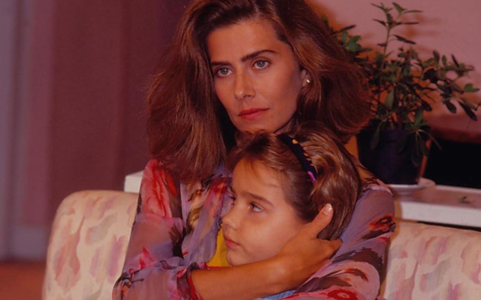 As atrizes Maitê Proença e Tatyane Goulart em cena da novela Felicidade; Maitê abraça a cabeça de Tatyane, ainda criança