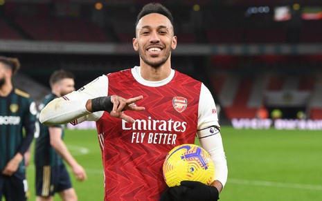 Aubameyang sorri com uniforme vermelho do Arsenal, com mangas brancas, e luva preta na mão esquerda com a qual segura a bola, com a mão direita faz um sinal com três dedos