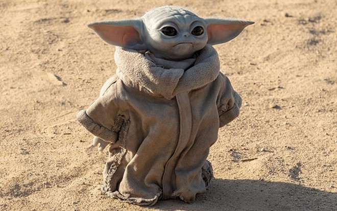 Baby Yoda veste uma roupinha cinza e caminha em um deserto em cena da série The Mandalorian