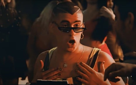 Trecho de videoclipe mostra o cantor Bad Bunny sentado, usando regata, óculos escuros posicionados no meio do nariz, cabelo raspado e fazendo uma expressão de surpresa