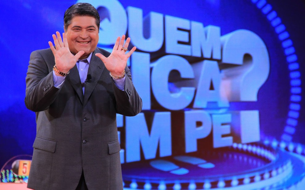 O apresentador José Luiz Datena usa terno escuro e camisa ao posar com palmas das mãos abertas no cenário do programa Quem Fica em Pé em 2012