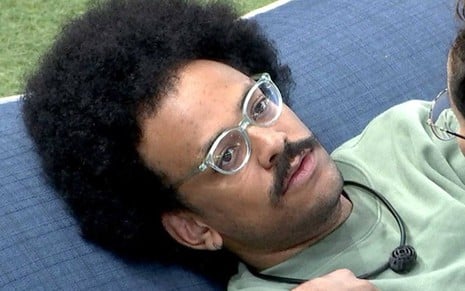 João está deitado em um sofá azul, usa camiseta verde, está com óculos de grau e olha para cima