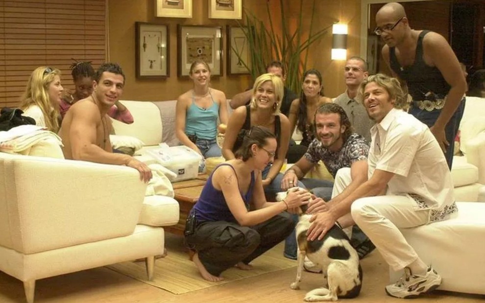 Participantes do Big Brother Brasil em 2002 brincam com cadela Molly na sala do programa, com sofás brancos
