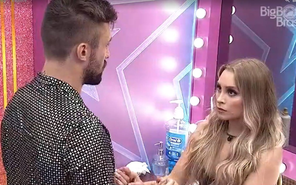 Arthur olha para Carla, ele usa camiseta cinza; Carla olha para Arthur, está com o cabelo solto e usa top preto