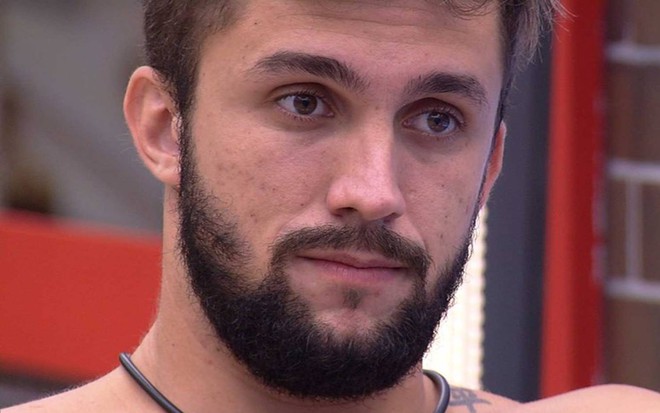 Arthur olha para o lado, está sem camiseta e está em frente a um espelho com moldura vermelha
