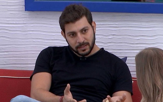 Caio olha para Carla, ele usa camiseta preta e está sentado em um sofá vermelho