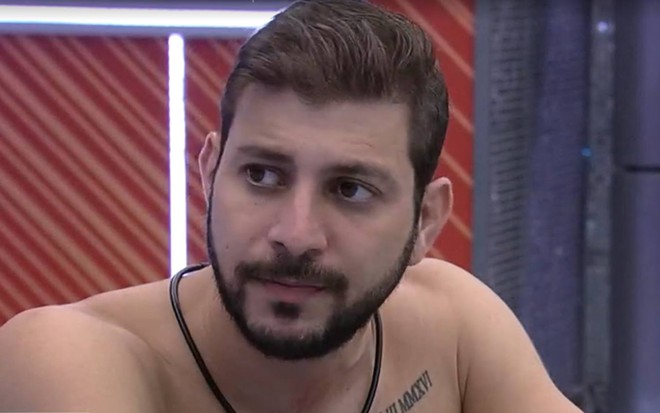 Caio olha para o lado, está sem camiseta e está na frente de um fundo vermelho